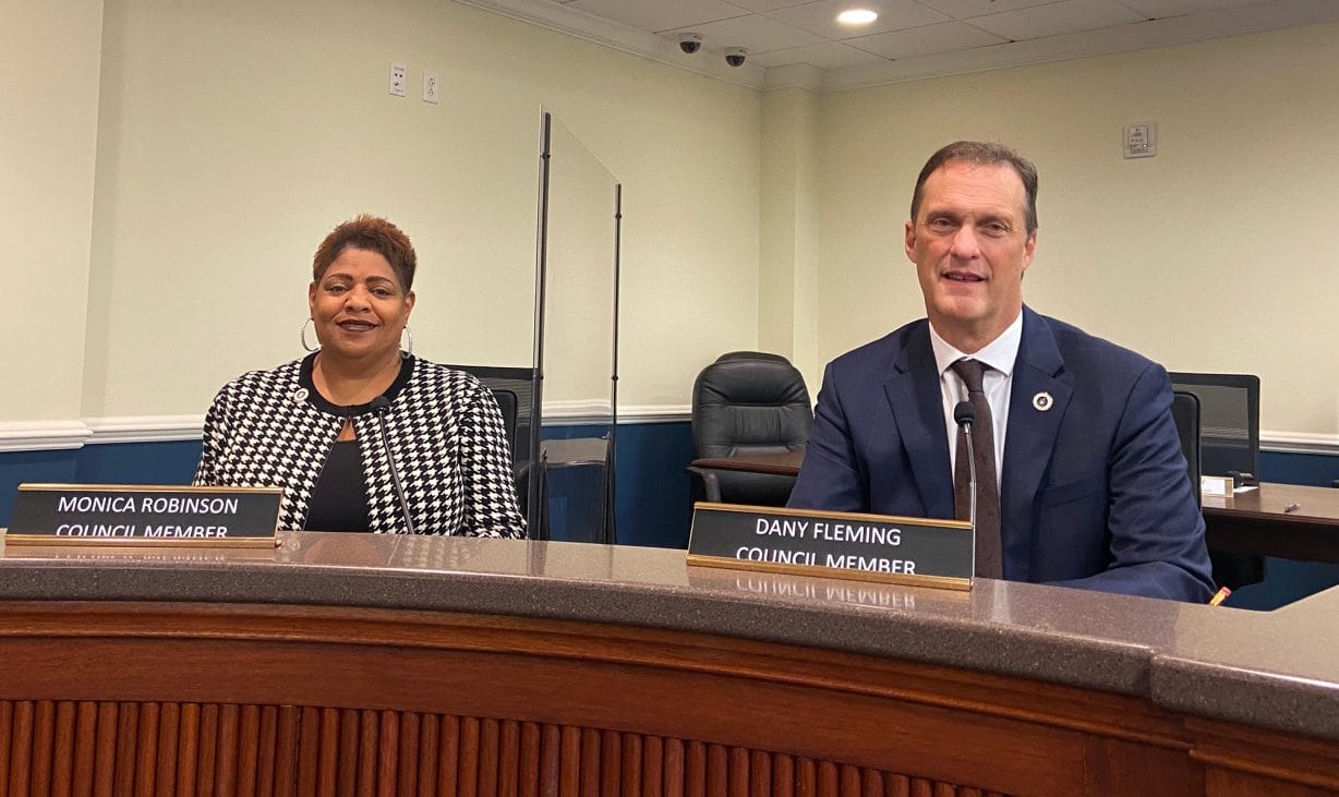 Two people sitting at the council dais look at the camera. 