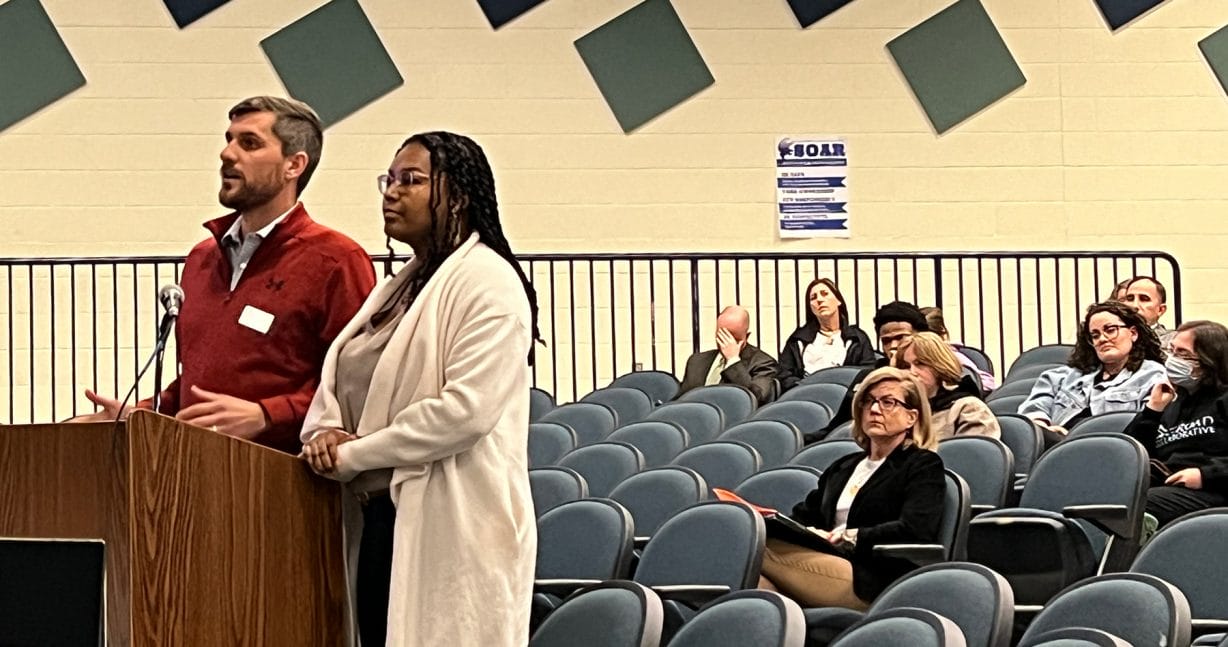 Two people stand at a podium while others sit in chairs behind them