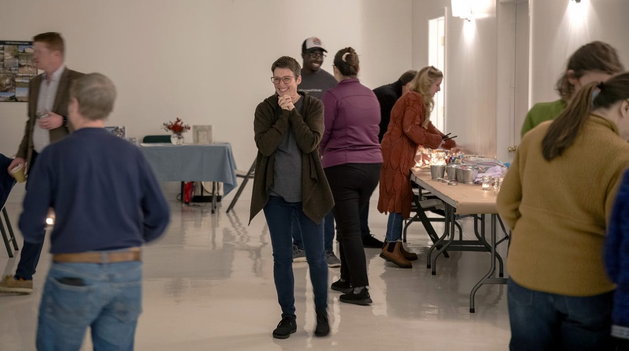 A woman clasps her hands in joy in the center of busy room as people talk to each other.