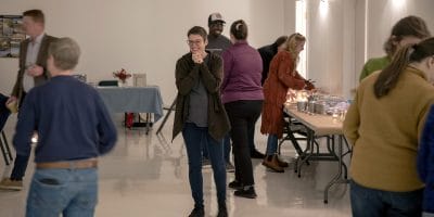 A woman clasps her hands in joy in the center of busy room as people talk to each other.