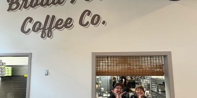 Two people in the opening from a kitchen under a painted sign that says Broad Porch Coffee Co.