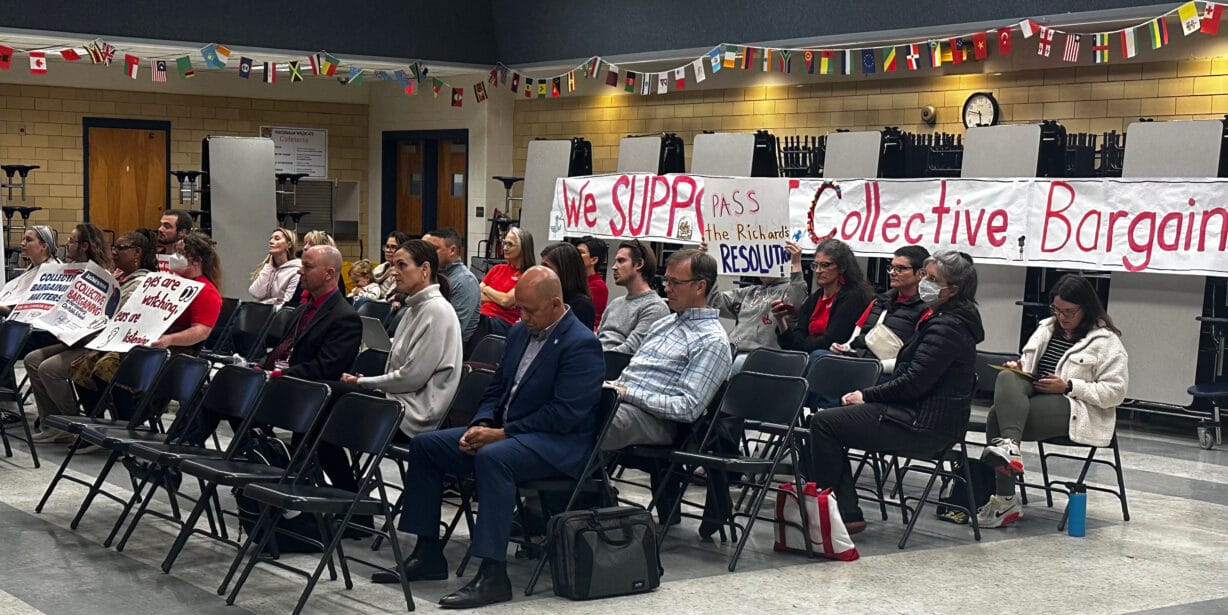 People sitting on folding chairs in a school cafeteria in front of a banner reading "We Support Collective Bargaining"