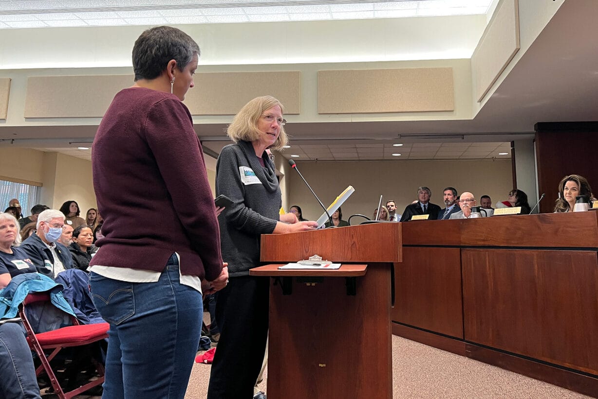 Two women at a podium speak to a group sitting on a dais