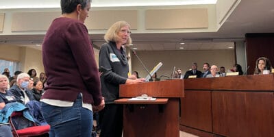 Two women at a podium speak to a group sitting on a dais