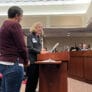 Two women at a podium speak to a group sitting on a dais