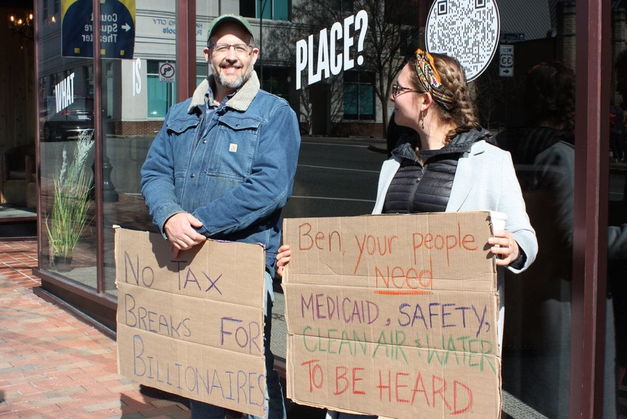 A man holds a cardboard sign that says "No tax breaks for billionaires," and. woman holds a sign that says: "Ben, your people need Medicaid, safety, clean air & water to be heard." 