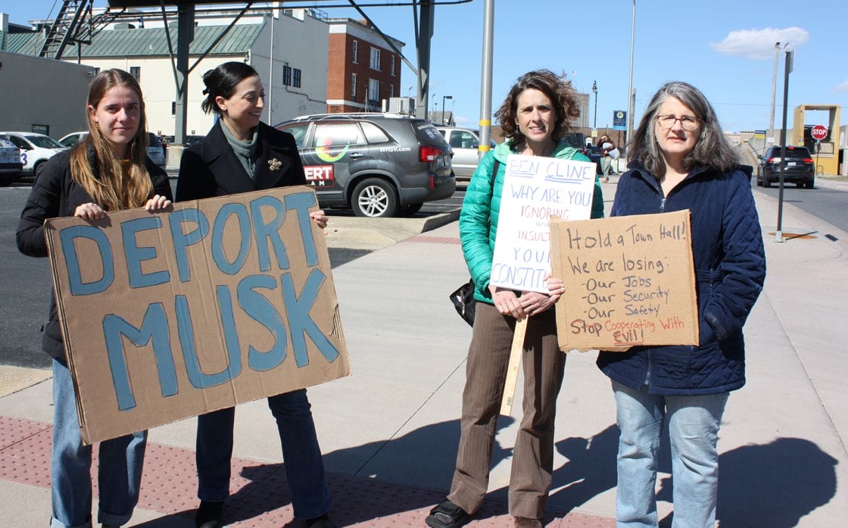 Four women holding hand-drawn signs on cardboard. One says "Deport Musk." The other call on Cline to hold a town hall meeting. 