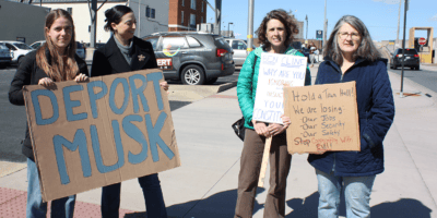 Four women holding hand-drawn signs on cardboard. One says "Deport Musk." The other call on Cline to hold a town hall meeting.