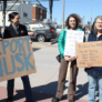 Four women holding hand-drawn signs on cardboard. One says "Deport Musk." The other call on Cline to hold a town hall meeting.