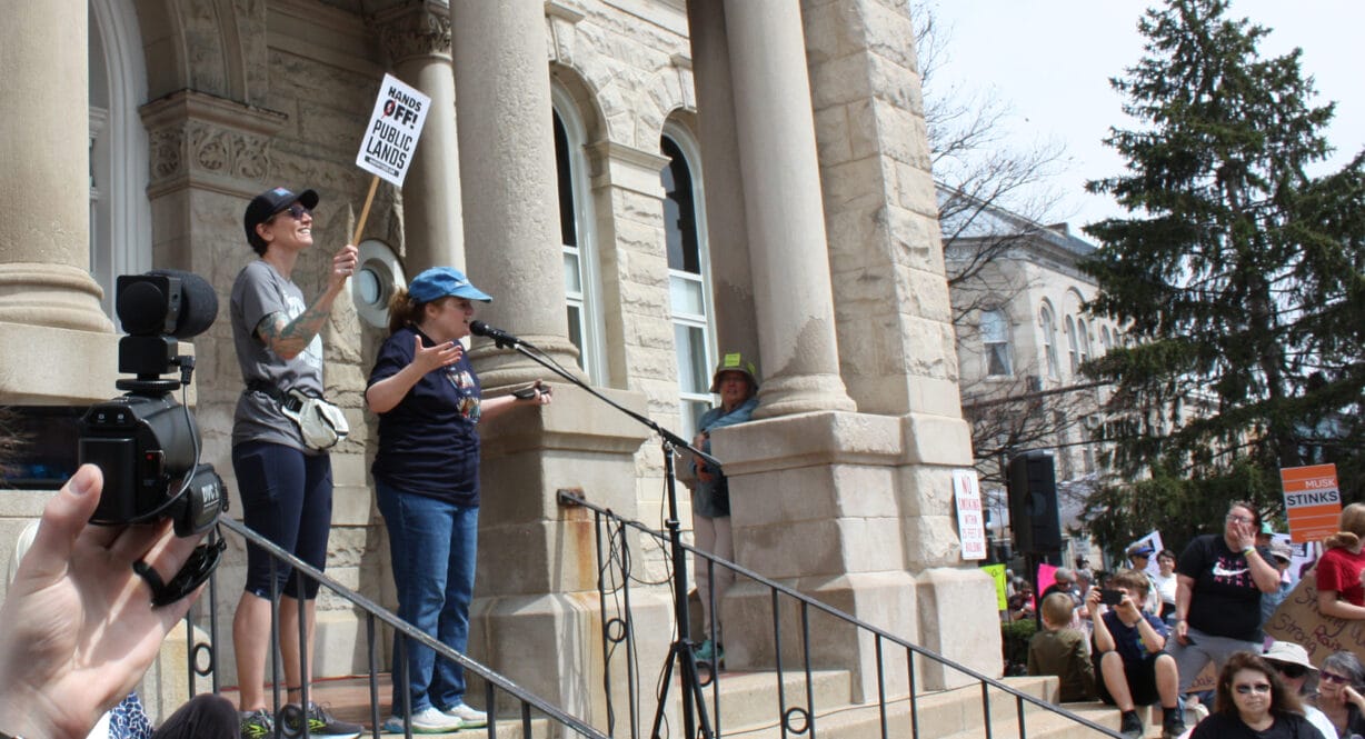A woman holding a sign and a woman at a microphone address a crowd
