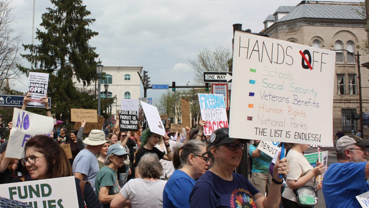 A crowd of people holding signs, including one that says "Hands Off" and lists federal agencies and programs