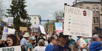 A crowd of people holding signs, including one that says "Hands Off" and lists federal agencies and programs