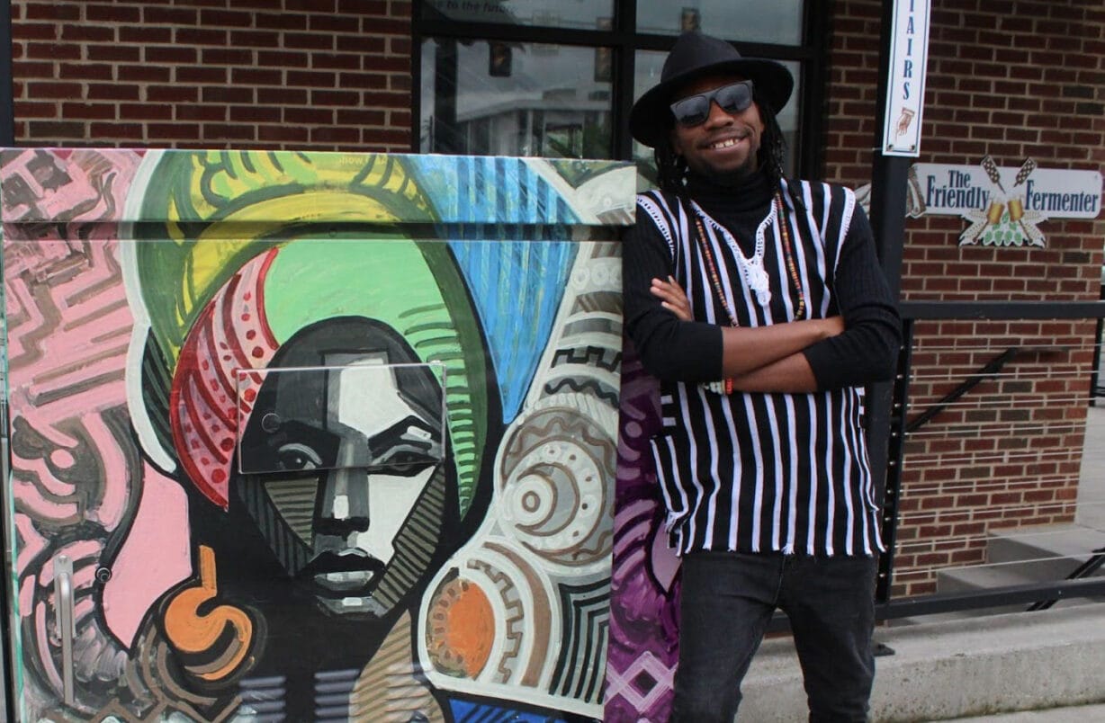 A man stands with his arms crossed and smiling next to a mural of a colorfully dressed woman