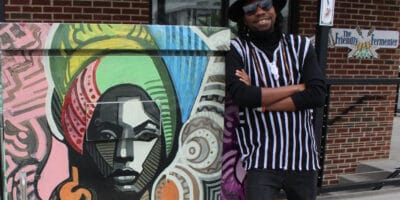 A man stands with his arms crossed and smiling next to a mural of a colorfully dressed woman