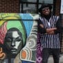 A man stands with his arms crossed and smiling next to a mural of a colorfully dressed woman