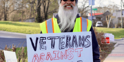 A man in a "Veterans" baseball cap and yellow safety vest holds a homemade sign saying "Veterans against Trump"