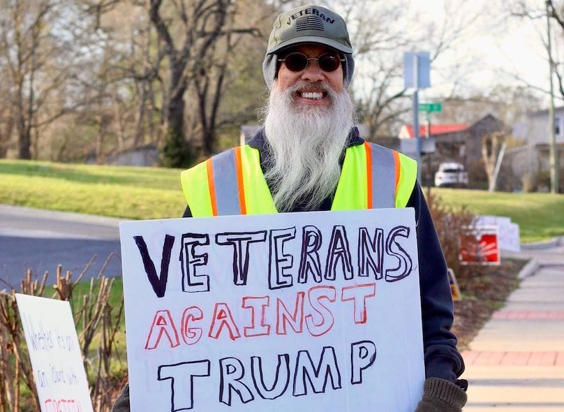 A man in a "Veterans" baseball cap and yellow safety vest holds a homemade sign saying "Veterans against Trump"